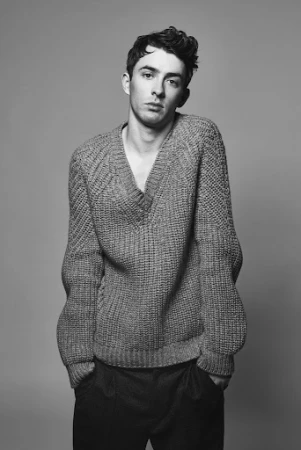 Black and white portrait of a young adult man wearing a wool V-neck sweater and dark pants in front of a neutral grey background.