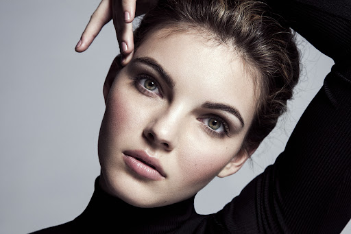 Close-up portrait of a woman in a black turtleneck with hands raised near her head, against a neutral grey studio background.