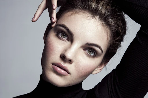 Close-up portrait of a woman in a black turtleneck with hands raised near her head, against a neutral grey studio background.