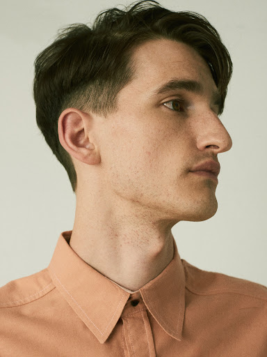 Portrait of a young adult man with dark hair in a salmon-colored collared shirt against a neutral background, looking off to the side.