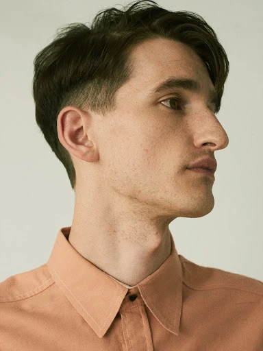 Portrait of a young adult man with dark hair in a salmon-colored collared shirt against a neutral background, looking off to the side.