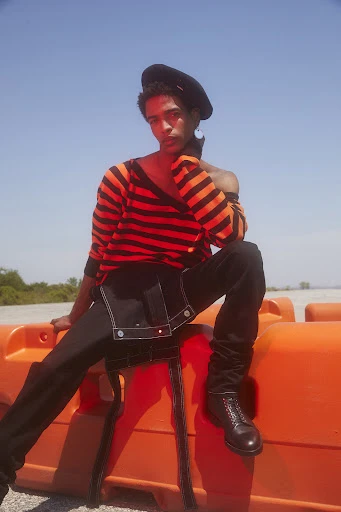 A male model poses outdoors, wearing a striped sweater, dark pants, black boots, and a beret, sitting on an orange barrier under a blue sky.