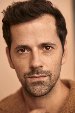 Close-up portrait of a man in his 30s with dark hair, light facial hair, and freckles against a neutral background.