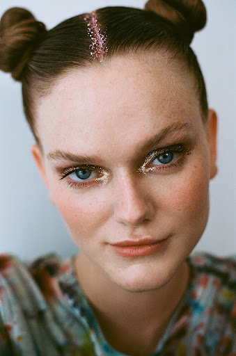Close-up of a young woman with creative gold eye makeup and pink glitter hair parting, styled with space buns against a white background.