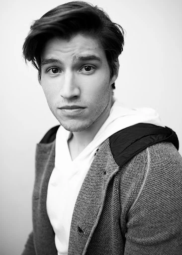 Black and white portrait of a young adult male with dark hair, wearing a white hoodie and a gray cardigan. Close-up shot, eye-level angle.