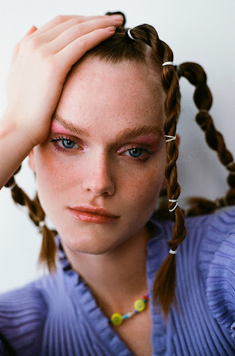 Close-up studio shot of a fair-skinned female model with red hair styled in braids and pink eye shadow wearing a periwinkle top.
