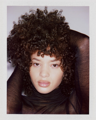 Portrait of a woman with voluminous dark curly hair wearing a black sheer top against a white backdrop.