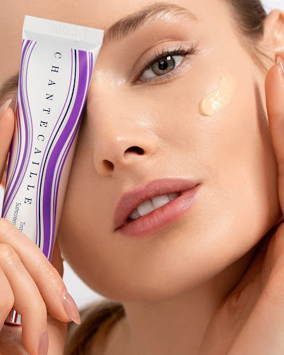 Close-up of a woman with fair skin and a drop of foundation on her cheek, holding a Chantecaille skincare tube.