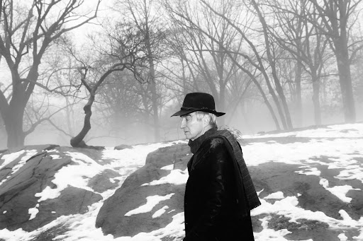 Elderly man in a dark fedora and coat walking through a snowy, misty park with bare trees.