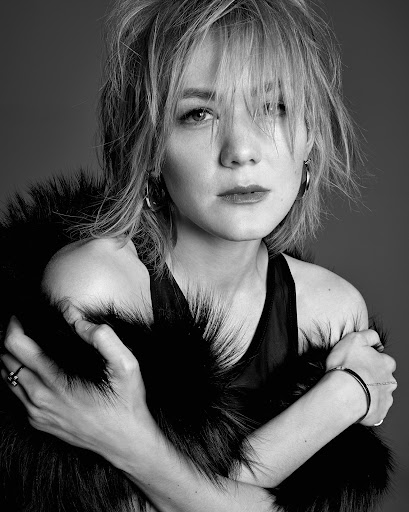 Black and white portrait of a blonde woman with a fur shawl, wearing a black top and hoop earrings, with her arms folded.