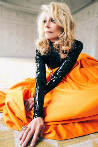 Elegant older woman with blonde hair poses in an orange dress and black sequin top indoors, creating a striking fashion portrait.