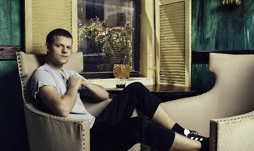 Man sitting in an upholstered chair indoors, wearing a light-gray T-shirt and black pants. A window with shutters is behind him.