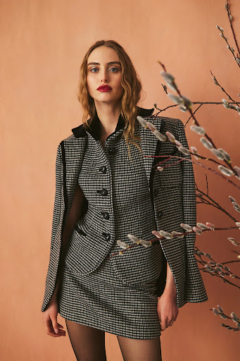Elegant woman in a houndstooth skirt suit poses indoors near willow branches, showcasing a sophisticated fashion style with a neutral backdrop.