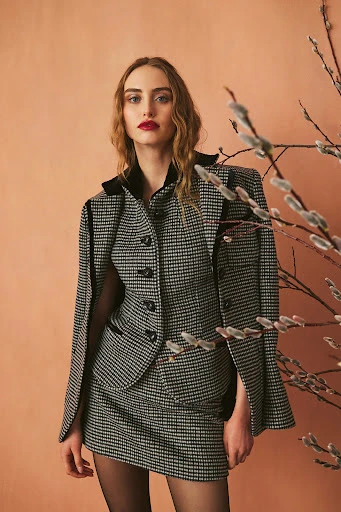 Elegant woman in a houndstooth skirt suit poses indoors near willow branches, showcasing a sophisticated fashion style with a neutral backdrop.
