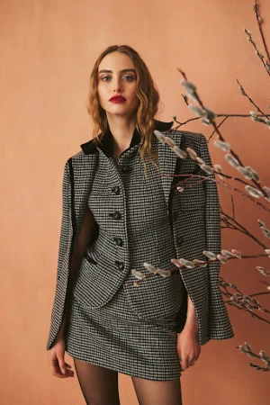 Elegant woman in a houndstooth skirt suit poses indoors near willow branches, showcasing a sophisticated fashion style with a neutral backdrop.