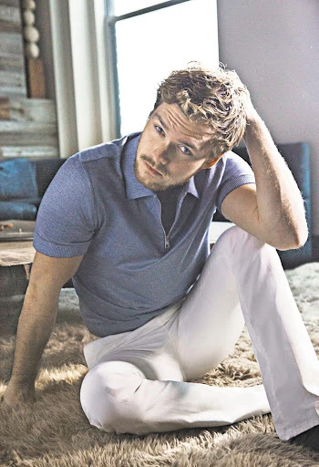 Young man with blond hair sitting on a tan shaggy carpet in natural light wearing white pants and a blue collared shirt.