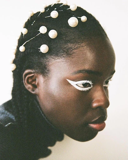 Close-up of a dark-skinned woman with pearls in her hair and white eyeliner looking off to the side, wearing a dark shirt.