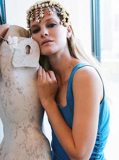 Adult woman in blue dress wearing pearl headpiece poses near mannequin in front of window. Interior fashion shot.