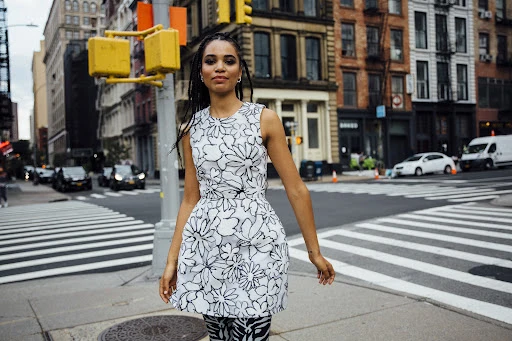 Young woman with braided hair in white dress with black floral pattern crossing a city street with buildings and cars in the background.