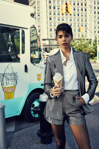 Adult female fashion model in a glittery short suit holds an ice cream cone on a city sidewalk next to an ice cream truck.