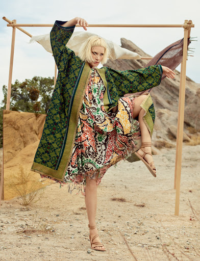 A model poses outdoors wearing a green and brown patterned kimono-style jacket over a multicolored floral dress and tan strappy sandals.
