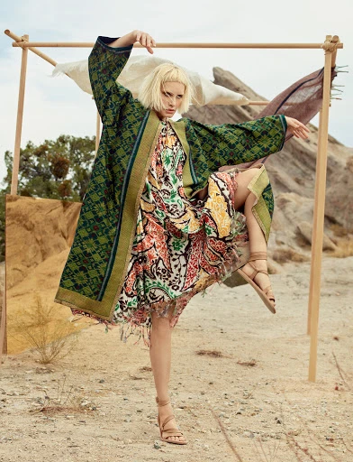 A model poses outdoors wearing a green and brown patterned kimono-style jacket over a multicolored floral dress and tan strappy sandals.