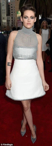 Kristen Stewart poses on a red carpet wearing a white dress and gray high heels, with a woman in the background.