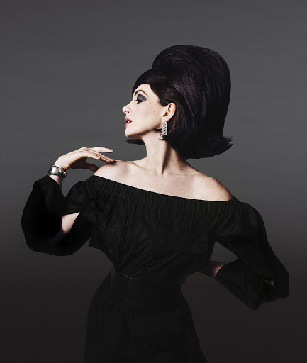 Elegant woman with bouffant hairstyle, off-shoulder black dress, silver bracelet, and diamond earrings poses against a gray backdrop.