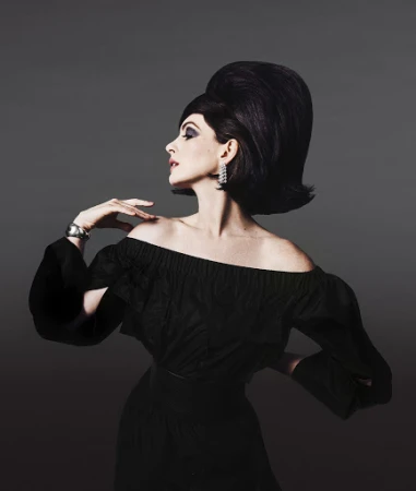 Elegant woman with bouffant hairstyle, off-shoulder black dress, silver bracelet, and diamond earrings poses against a gray backdrop.