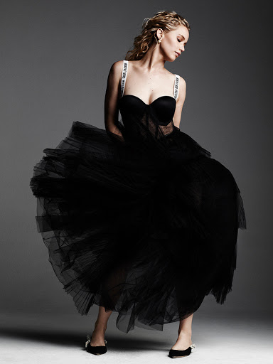 Fashionable blonde woman poses gracefully in a black tulle dress with logo straps in a studio shot with a neutral gray background.