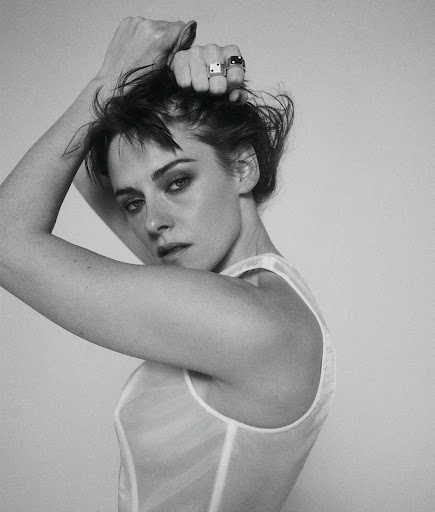 Monochrome portrait of a woman with short, dark hair, posing in a sheer white top with hands near her head, against a plain background.