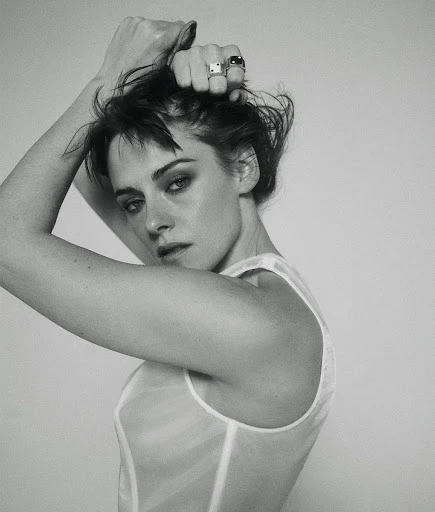 Monochrome portrait of a woman with short, dark hair, posing in a sheer white top with hands near her head, against a plain background.