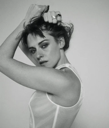 Monochrome portrait of a woman with short, dark hair, posing in a sheer white top with hands near her head, against a plain background.