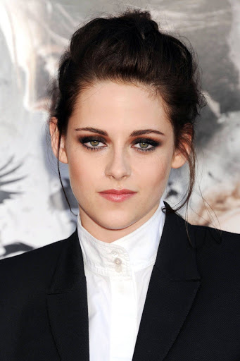 Headshot of Kristen Stewart at event in a black suit and white collared shirt, her hair in an updo, posing in front of an out-of-focus background.