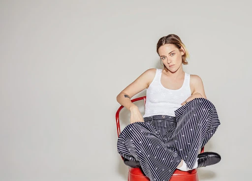 Kristen Stewart seated cross-legged on a bright red chair wearing a white tank top and patterned navy trousers against a plain light grey wall.
