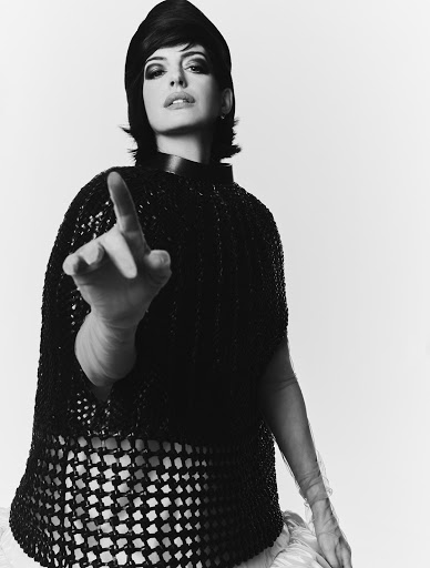 Monochrome portrait of a stylish woman with retro-inspired hair and makeup, wearing a net cape, with her finger pointing forward on a white background.