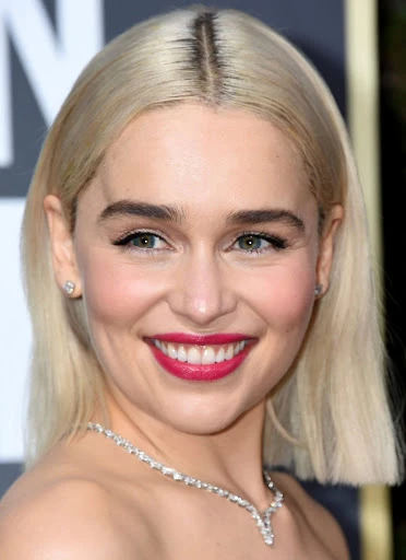 Close-up portrait of a fair-skinned woman with blonde bob hair, smiling with red lipstick, wearing a diamond necklace, at an indoor event.