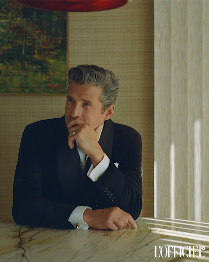 Portrait of Patrick Dempsey in black tuxedo at marble table with hand to face in studio with L'Officiel magazine logo in foreground.