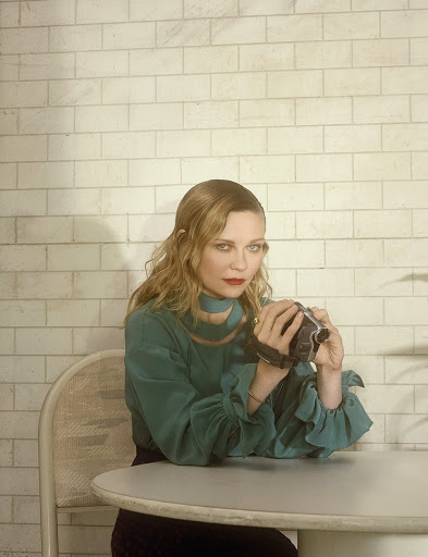 A woman with wavy blonde hair is seated at a table, holding a camcorder. She's wearing a teal blouse and the background is a tiled wall.