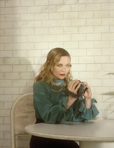 A woman with wavy blonde hair is seated at a table, holding a camcorder. She's wearing a teal blouse and the background is a tiled wall.