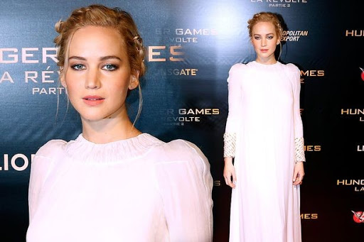 Jennifer Lawrence poses in a white gown at a premiere event with a logo backdrop featuring movie title. Close-up and full shot included.