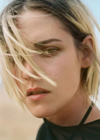 Close-up portrait of a woman with blonde hair blowing across her face and dark clothing against an outdoor sky background