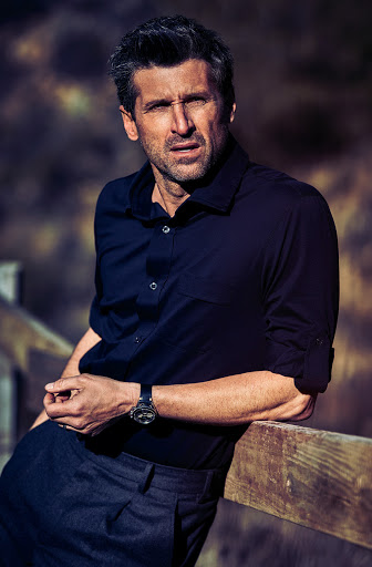 Portrait of adult male outdoors wearing a black button-down shirt and leaning against a wooden fence with a blurred background.
