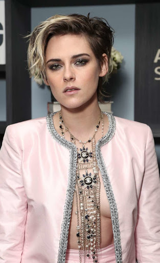 Close-up of Kristen Stewart wearing a pink Chanel jacket and multiple necklaces, posing indoors against a neutral-toned wall with shelves.