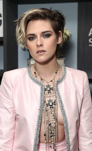 Close-up of Kristen Stewart wearing a pink Chanel jacket and multiple necklaces, posing indoors against a neutral-toned wall with shelves.