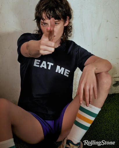 Adult woman in a black T-shirt with 'EAT ME' pointing to the camera while sitting indoors with a concrete wall background.