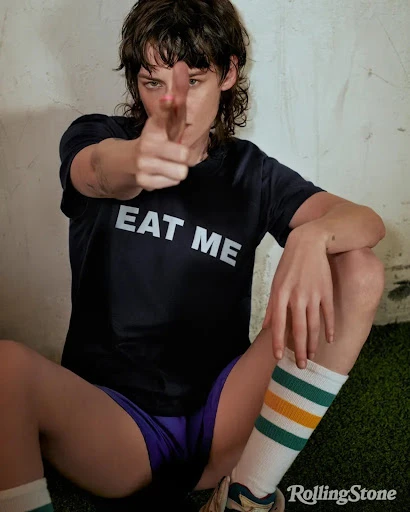 Adult woman in a black T-shirt with 'EAT ME' pointing to the camera while sitting indoors with a concrete wall background.
