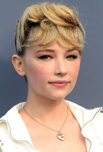 Portrait of Haley Bennett with blonde pinned-up hair, winged eyeliner, and a heart locket necklace against a grey backdrop.