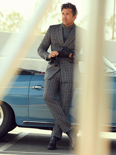 Adult male in a grey plaid suit poses next to a vintage blue convertible car in a stylish outdoor automotive fashion shot.