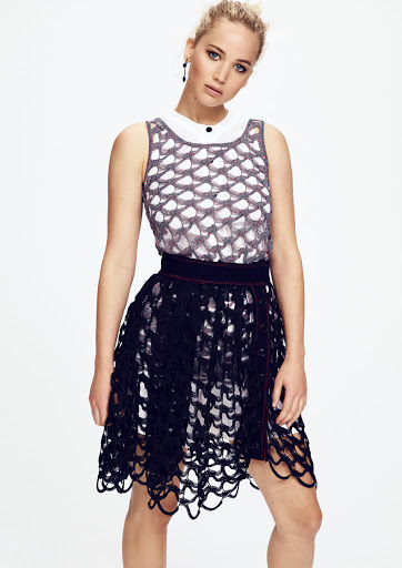 Jennifer Lawrence poses in a fashion shot wearing a grey and white top and black skirt against a bright white background.